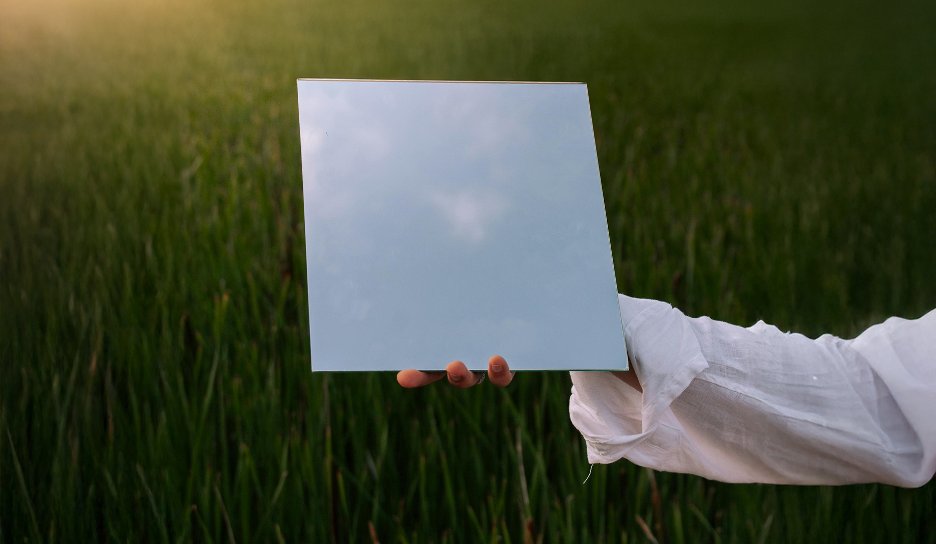 Hand holding a mirror reflecting the sky, set against a grassy field